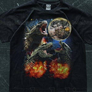 以下が含まれることがあります： 黒いTシャツ。ゴジラのような3匹の生き物が咆哮するグラフィック、背景に惑星があります。デザインには炎と稲妻が含まれており、ドラマチックなシーンを作り出しています。