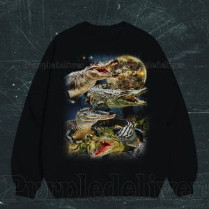 May include: Black sweatshirt featuring a detailed graphic of several alligators with open mouths, set against a backdrop of a full moon and starry sky. The design showcases the alligators in various poses, highlighting their textures and colors.
