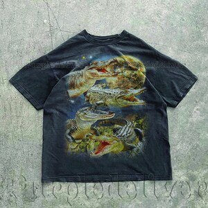 May include: Dark gray t-shirt featuring a detailed graphic of several alligators with open mouths, set against a night sky and moon backdrop. The design showcases the alligators in shades of brown, green, and white.