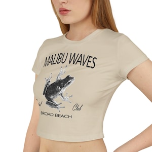 Könnte beinhalten: Beiges Crop-Top mit kurzen Ärmeln, auf dem der Text "MALIBU WAVES" über einer schwarzen Frosch-Illustration steht. Darunter stehen die Worte "Social Club" und "BROAD BEACH".
