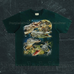 May include: Dark green t-shirt featuring a detailed graphic of several alligators with open mouths, set against a night sky and a full moon. The design includes various shades of green, brown, and white, creating a striking contrast on the shirt.
