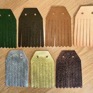 May include: A collection of leather fringe keychains in various colors, including green, brown, tan, silver, yellow, and dark brown. Each keychain has a rectangular top with two holes and a fringe bottom. The keychains are arranged on a wooden surface.