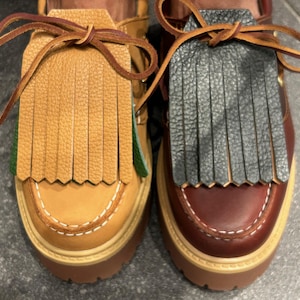 May include: A pair of leather loafers with fringed accents. One shoe is tan with a light brown fringe, the other is burgundy with a gray fringe. Both have thick, brown soles and decorative stitching.