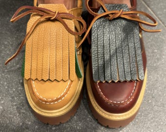 May include: A pair of leather loafers with fringed accents. One shoe is tan with a light brown fringe, the other is burgundy with a gray fringe. Both have thick, brown soles and decorative stitching.
