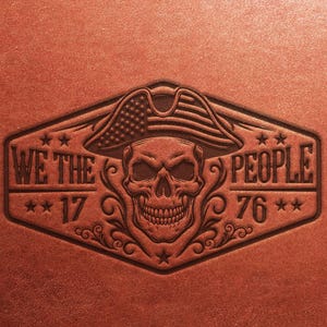 Puede incluir: Un parche de cuero marrón con una calavera que lleva un sombrero tricornio con el diseño de la bandera estadounidense. El texto "WE THE PEOPLE 1776" está grabado en relieve alrededor de la calavera, con estrellas y adornos decorativos.