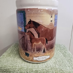 May include: A white ceramic jar with a painted scene of a brown horse and foal in a barn. The jar has a rectangular label that reads "Barn Buddies by Persia Clayton Weir". A cat is visible in the background.