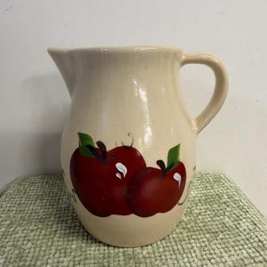 Puede incluir: Jarra de cerámica color crema con asa y boquilla. La jarra está decorada con dos manzanas rojas con hojas verdes. La jarra está sobre una superficie verde texturizada.
