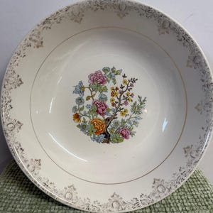 May include: Off-white ceramic serving bowl with a floral design in the center. The floral design features pink, orange, and yellow flowers with green and blue leaves. The rim has a gold trim and a decorative pattern.