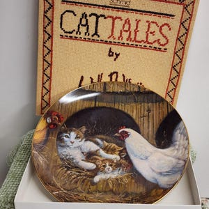 May include: A decorative plate featuring a cat family nestled in a barn setting with a white hen. The plate has a gold rim and is set against a backdrop of a stitched fabric panel with the words "CATTALES" in red and black.