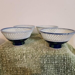 May include: Four white ceramic bowls with a lattice design. Each bowl has a blue rim and sits on a dark blue base with green floral accents. The bowls are arranged on a textured green surface.