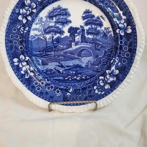 May include: Blue and white decorative plate with a landscape scene. The plate has a scalloped edge with floral designs. The centre of the plate features a bridge, trees, and a building. The plate is displayed on a gold-coloured stand.