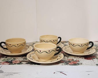 May include: A set of four cream-coloured teacups and saucers. Each cup has a black handle, a green wavy line, and small red dots. The saucers have a matching design. The set is on a floral patterned surface.