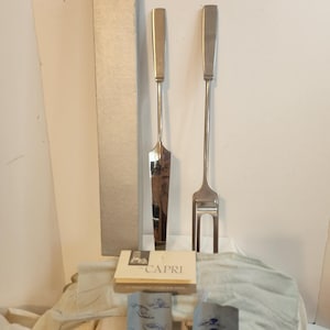 May include: Silver-toned serving set, including a cake server and a fork, with rectangular handles. The set is displayed with a box and a card that reads "CAPRI". The items are on a light-colored surface.