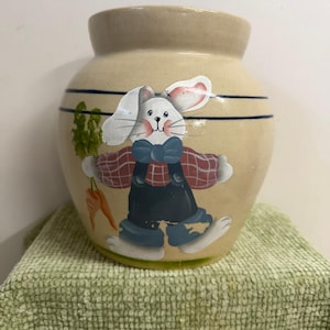 May include: A beige ceramic jar with a painted Easter bunny design. The bunny wears a plaid shirt, blue overalls, and a blue bow tie. The jar has two blue stripes and a green carrot and plant design.