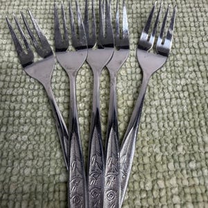 Könnte beinhalten: Fünf Edelstahlgabeln mit einem dekorativen Muster auf den Griffen. Die Gabeln sind fächerförmig angeordnet, wobei die Zinken nach oben zeigen. Die Griffe haben ein geprägtes Design. Die Gabeln liegen auf einer strukturierten grünen Oberfläche.