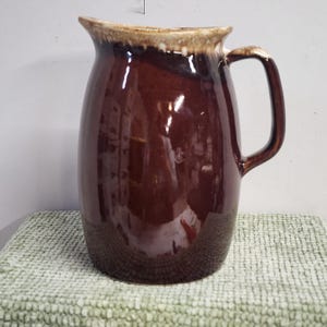 May include: A brown ceramic jug with a handle. The jug has a glossy finish and a lighter-coloured rim. The jug is likely used for serving beverages or as a decorative item.