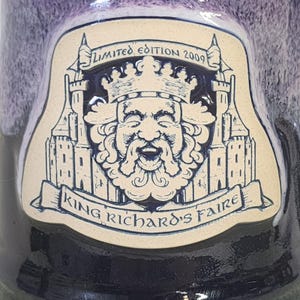 May include: A ceramic mug with a decorative emblem featuring a crowned king's face, castles, and the text "King Richard's Faire" and "Limited Edition 2009". The mug has a dark blue and purple glaze.