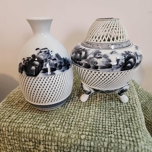 Puede incluir: Dos jarrones de porcelana blancos y azules. Uno es un jarrón en forma de botella con un diseño de celosía en la mitad inferior y una pintura de paisaje. El otro es un jarrón redondo con un diseño de celosía y tres patas pequeñas.
