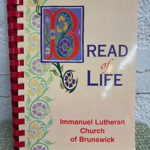 Puede incluir: Un libro de espiral con una cubierta beige que presenta una letra B roja grande y ornamentada decorada con patrones florales coloridos. El texto dice BREAD of LIFE e Immanuel Lutheran Church of Brunswick en letras rojas y azules.
