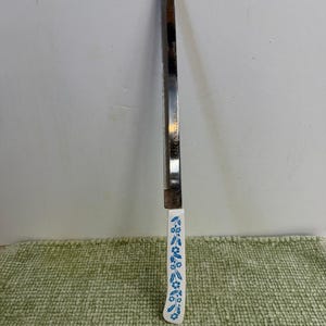 Puede incluir: Un cuchillo de pan de acero inoxidable con un mango blanco con un diseño floral azul. El cuchillo se muestra verticalmente sobre un fondo blanco y una superficie verde texturizada.