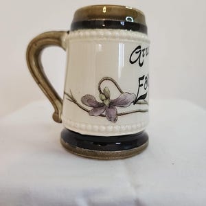 May include: A small ceramic mug with a handle. The mug is cream-colored with a brown top and base. It features a purple flower design and the words "Grandpa's Favorite" in black script.