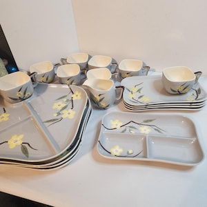 Puede incluir: Un juego de vajilla vintage de cerámica gris con diseños florales amarillos. El juego incluye tazas, platillos, bandejas para servir y platos divididos. Las tazas y las piezas para servir tienen asas y bordes marrones. El diseño floral presenta flores amarillas y hojas verdes.