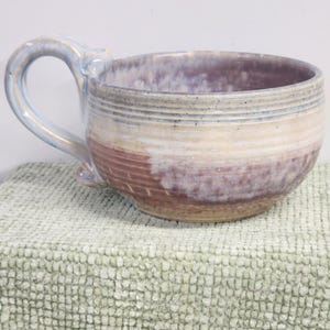 May include: A handmade ceramic mug with a large handle. The mug features horizontal bands of color, including shades of purple, beige, and blue. The mug is resting on a textured, light green surface.