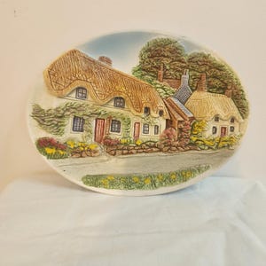 Puede incluir: Plato decorativo ovalado de cerámica con un relieve detallado de casas con tejados de paja. El plato tiene un fondo de cielo azul claro, árboles verdes y una carretera. Las casas tienen puertas y ventanas rojas.