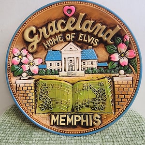 May include: A round Graceland souvenir plate. The plate features a depiction of Elvis's home, with the text "Graceland HOME OF ELVIS" in gold lettering. Pink and white flowers and musical notes are also included. The word "MEMPHIS" is at the bottom.
