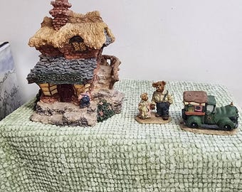 May include: A collection of miniature bear figurines and a shop. The shop has a thatched roof and the sign reads "TED E BEAR SHOP". Two bear figurines stand near a green truck. The figurines are on a green textured surface.