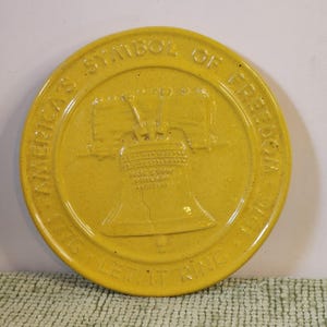 May include: Yellow ceramic plate with embossed text and image of the Liberty Bell. The plate reads "America's Symbol of Freedom" and "Let it Ring 1776". The plate is round and has a slightly textured surface.