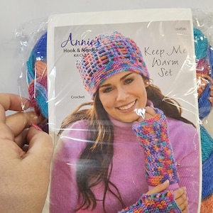 Könnte beinhalten: Ein Häkelset von Annie's Hook & Needle Kit Club mit einem "Keep Me Warm Set". Das Set beinhaltet eine Mütze und fingerlose Handschuhe in einem farbenfrohen, offen gewebten Design. Das Set ist mit Garn in Blau-, Rosa- und Orangetönen verpackt.