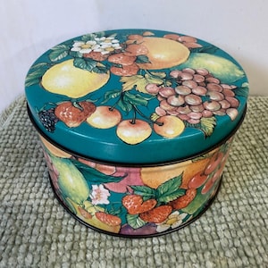 May include: A round, teal-colored tin with a lid, decorated with a vibrant fruit design. The artwork includes lemons, strawberries, grapes, and other fruits. The tin has a black rim and sits on a textured, light green surface.