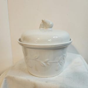 Op de afbeelding: Een witte keramische container met deksel en peervormige handgreep. De container heeft een ronde basis en een deksel. De body van de container is versierd met een blad- en wijnstokpatroon. De container staat op een wit oppervlak.