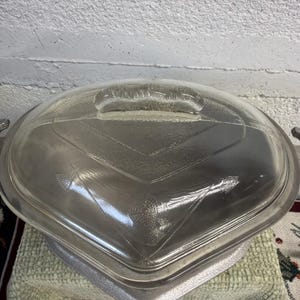 Puede incluir: Una fuente de vidrio transparente para horno con tapa a juego, que presenta un patrón geométrico en relieve en la superficie superior. La tapa tiene un asa central y la fuente incluye asas laterales, mostrando un diseño transparente con texturas.