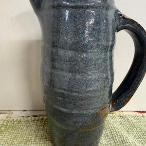 May include: A handcrafted ceramic jug with a dark blue glaze. It features a large handle and a pouring spout. The surface has a textured, organic look. This jug is suitable for serving water, juice, or other drinks. It is a unique piece of pottery.