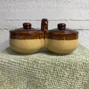 May include: A ceramic double condiment server with two lidded compartments and a handle. The server is two-toned, with a light tan base and a dark brown top and lid. The server is sitting on a green textured surface.