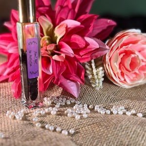 Puede incluir: Un frasco de perfume rectangular transparente con tapa dorada, que contiene un líquido oscuro, se encuentra frente a una vibrante flor rosa y una rosa color melocotón. Perlas blancas dispersas sobre una superficie de arpillera.