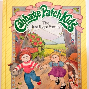 May include: A yellow book cover with a green and pink Cabbage Patch Kids logo. The title is "The Just-Right Family". The cover features an illustration of two Cabbage Patch Kids dolls walking in a forest with a squirrel and a bird.