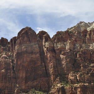 Może przedstawiać: Zdjęcie wysokiej formacji skalnej z czerwonobrązowymi klifami pod częściowo zachmurzonym błękitnym niebem. Ściana skalna ma pionowe prążki i różne odcienie brązu i beżu. Obraz przedstawia naturalny krajobraz.