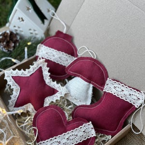 May include: A collection of burgundy Christmas ornaments in various shapes, including a star, heart, and stocking, with white lace trim. The ornaments are displayed in a cardboard box, suggesting a festive theme.
