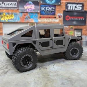 Puede incluir: Un modelo de juguete gris de un Hummer H1, con grandes neum&aacute;ticos negros todoterreno y un exterior detallado. El veh&iacute;culo en miniatura tiene techo r&iacute;gido y ventanas visibles, con luces traseras rojas. El modelo est&aacute; sobre un fondo de grafitis y ladrillos.