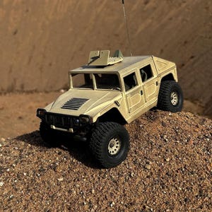 Puede incluir: Humvee de juguete de color beige con neum&aacute;ticos negros y una parrilla delantera negra. El veh&iacute;culo en miniatura tiene un dise&ntilde;o detallado, que incluye una estructura montada en el techo y una antena de radio. El juguete est&aacute; colocado sobre un peque&ntilde;o mont&oacute;n de arena.