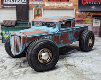Puede incluir: Un modelo de camioneta azul y óxido desgastado, con neumáticos negros grandes. El camión tiene un parabrisas agrietado y una parrilla delantera detallada. El fondo presenta grafitis y letreros.
