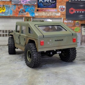 Puede incluir: Modelo Hummer verde oliva con neum&aacute;ticos grandes y robustos. La parte trasera del veh&iacute;culo muestra la palabra "HUMMER" y las letras "TTS". El modelo tiene una ventana trasera transparente y orificios de bala simulados.