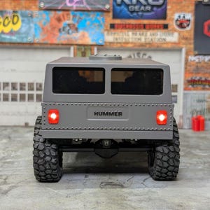 Puede incluir: Un modelo gris de Hummer con ventanas negras y neum&aacute;ticos grandes y texturizados. La palabra "HUMMER" se muestra en la parte trasera. Luces rojas son visibles en las esquinas traseras del veh&iacute;culo. El modelo est&aacute; sobre un fondo de letreros coloridos.