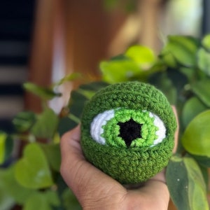May include: A green crocheted eye with a white iris and black pupil. The eye is being held in a hand.