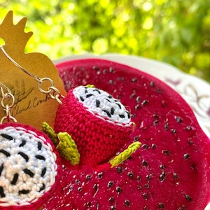 May include: Crocheted dragon fruit earrings with white and black details, displayed on a slice of fresh dragon fruit. The earrings are attached to a silver hoop.