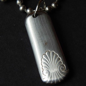 May include: A silver necklace with a rectangular pendant featuring an engraved design of a stylized seashell.