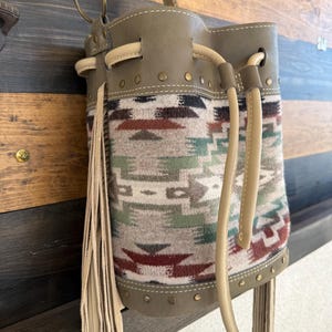 May include: A tan leather bucket bag with a Southwestern-style patterned fabric insert. The bag features a drawstring closure, studded accents, and a long fringe detail. The bag is suspended from a brass-colored ring.